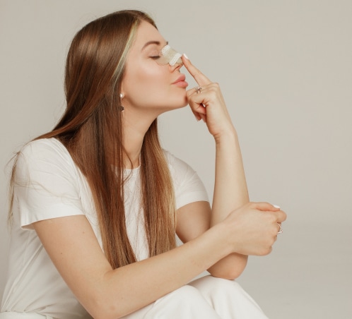 Side profile of a woman gently touching a small adhesive bandage on her nose, suggesting post-operative care after rhinoplasty, against a neutral background.
