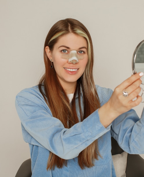 Smiling woman with a nasal bandage holding a mirror, checking her results after a nose job in Turkey, highlighting post-operative rhinoplasty recovery.